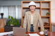 © Ekkasit A Siam - Asian female designer standing at a wooden desk, smiling with a white hard hat. Casual attire in a modern office setting with a laptop, coffee cup, Positive and confident expression.