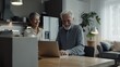 © The Little Hut - Happy mature older couple using laptop technology at home Smiling middle aged senior man working on computer sitting at table with wife standing nearby in living room Vertical candid s : Generative AI