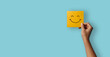 © KF - A hand holding a yellow happy smile face on blue background. Positive customer rating, good feedback and review. Happiness and world mental health day concept