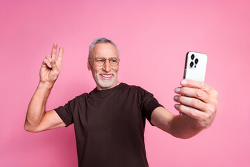 Sticker - Portrait of cheerful man with beard dressed t-shirt in glasses show v-sign make selfie on smartphone isolated on pink color background