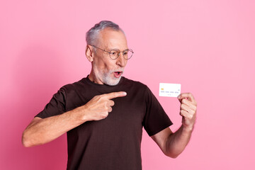 Wall Mural - Portrait of impressed senior man with beard dressed t-shirt in glasses indicating look at credit card isolated on pink color background