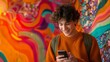 © Thanaphon - Young man using smartphone, smiling in front of vibrant, colorful urban street art mural. Casual modern lifestyle concept.