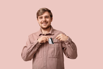 Wall Mural - Young smiling man putting credit card in shirt pocket on pink background