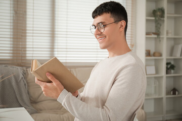 Wall Mural - Handsome happy young man in new stylish eyeglasses reading book at home