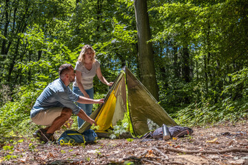 Fototapete - A couple sets up a tent together in nature on a sunny summer day. They work as a team, helping each other to prepare their campsite.