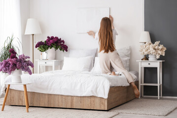 Poster - Young woman hanging picture on white wall near vases of lilacs in bedroom