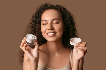 Wall Mural - Beautiful young African-American woman with jar of face cream on brown background