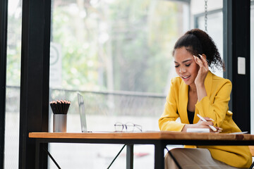 Wall Mural - A young woman in a yellow blazer works on her laptop in a bright, modern office with large windows and natural light.