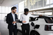 © sofiko14 - Dealer and businessman discussing features of electric SUV in car dealership. Businessman evaluates electric vehicle's specifications. Interaction focusing on transportation and modern technology.