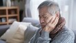 © KumCup - An elderly man sitting on a couch indoors, holding his cheek in pain and wearing a gray sweater and a brown scarf.