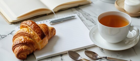 Canvas Print - Tea break concept with a setup featuring a tea cup, croissant, tea spoons, blank notebook paper, pen set on a marble surface with a copy space image.