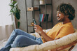 © ViDi Studio - Side view happy young Indian man wear orange casual clothes hold use mobile cell phone sits in armchair stay at home hotel flat rest relax spend free spare time in living room indoor. Lounge concept.