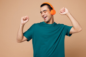 Wall Mural - Young smiling happy middle eastern man wear blue t-shirt casual clothes listen to music in headphones raise up hands dance isolated on plain pastel beige background studio portrait. Lifestyle concept.