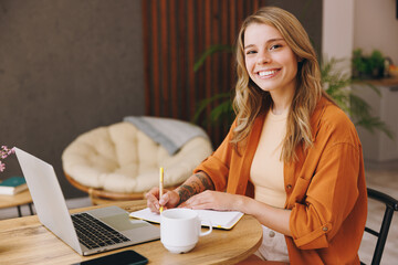 Wall Mural - Smiling young IT woman wear orange shirt hold use laptop pc computer write in notebook sit alone at table in coffee shop cafe restaurant indoor work or study. Freelance mobile office business concept.