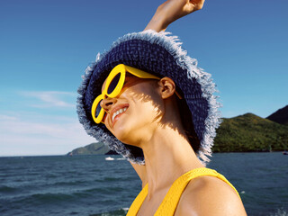 Beach scene with a woman in sunglasses and a hat, standing on the shore, ocean waves in the background, and distant mountains under a clear blue sky