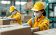 © praewpailyn - Two workers in yellow uniforms, wearing hard hats and masks, packing boxes in a busy warehouse environment, ensuring safety and efficiency.