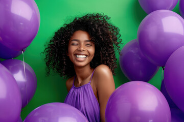 A beautiful young woman with curly hair smiles brightly amidst vibrant purple balloons and a green background