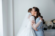 © Vasil - A bride and her bridesmaid are hugging each other. The bride is wearing a white dress and the bridesmaid is wearing a blue dress. Scene is happy and joyful
