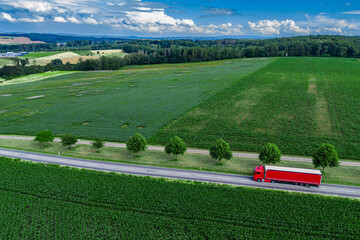 Wall Mural - Red cargo truck on the road. Asphalt road among green fields and beautiful clouds at sunset. Cargo delivery and transportation concept