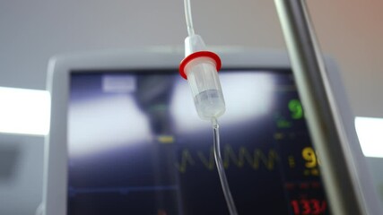 Wall Mural - Reservoir of iv drip with the medicine dripping inside. Low angle view. Screen of lung ventilator at backdrop in blur.