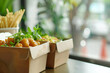 © Victor Bertrand - Freshly prepared take-out food in eco-friendly packaging, garnished with herbs, next to a serving of french fries on a wooden counter.