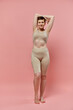 © LIGHTFIELD STUDIOS - A woman with short hair wearing a bra stands confidently, arms raised above her head, against a pink backdrop.