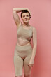 © LIGHTFIELD STUDIOS - A woman with short hair, wearing a beige sports bra, stands confidently in front of a pink background.