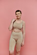 © LIGHTFIELD STUDIOS - A woman with short hair stands confidently in a beige bra and leggings against a pink background, promoting breast cancer awareness.