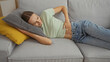 © Krakenimages.com - Young woman lying on a couch in a living room, appearing to have stomach pain, resting with a cushioned pillow.