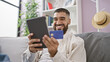 © Krakenimages.com - Smiling man with beard holding tablet and credit card indoors on sofa for online shopping or banking.