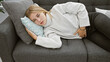 © Krakenimages.com - A pained blonde woman with blue eyes dressed in a white sweater resting on a couch indoors, holding her stomach.