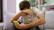 © Krakenimages.com - A young hispanic man with a beard looks distressed while using a smartphone in a modern living room setting.