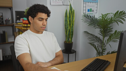 Poster - A handsome young hispanic man works attentively at his computer in a modern office, surrounded by plants and business charts, creating a professional and focused atmosphere.