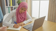 © Krakenimages.com - A focused woman with a hijab writing in a notebook by her laptop in a bright, modern home office setting.