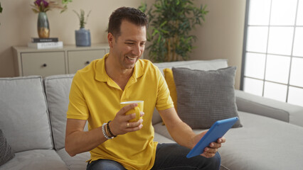 Middle-aged hispanic man in a yellow shirt enjoying coffee while using a tablet in a cozy living room