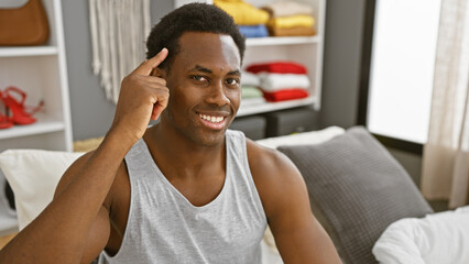 Wall Mural - Smiling young black man pointing at his head, sitting in a casual bedroom interior filled with light.