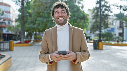 Wall Mural - Smiling hispanic man with beard using smartphone in sunlit city park.