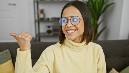 Wall Mural - A smiling young hispanic woman with glasses pointing sideways indoors at home.