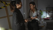 © Krakenimages.com - Two women investigating a crime scene indoors with one holding a phone and the other holding a notepad, surrounded by caution tape.