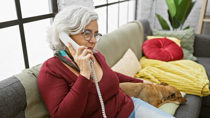 Wall Mural - Mature woman on phone with glasses, sitting with dog in cozy living room indoors.