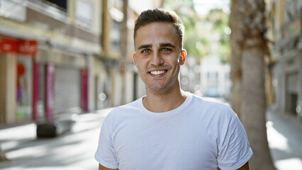 A smiling young hispanic man in casual attire posing confidently on a sunny urban street.