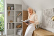 © Pixel-Shot - Mature woman reading magazine near hanging 3D painting on light wall at home