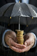 © Emanuel - Close-up of a businessman protecting a stack of coins with an umbrella