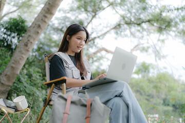 Wall Mural - Asian girl sit on a camping chair, working with computer notebook, relax on a relaxing day with nature