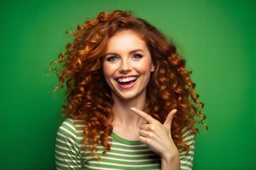 A beautiful red-haired, freckled, curly smiling girl on a green background shows with her hand.