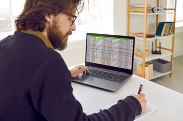 Wall Mural - Male entrepreneur or business accountant working on laptop computer. Bearded man sitting at office desk with notebook PC, working with database, using data sheets, writing down information in notepad