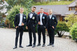 © Vasil - Four men in black suits and ties pose for a photo. The men are dressed in formal wear and are standing in front of a house. Scene is elegant and sophisticated