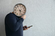 © Maxim Chuev - man in casual clothes holding round clock in front of his face, representing concept of time management. time is money idea. male  cover face, isolated on white wall background. mobile phone in hand