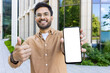 © Liubomir - Smiling man in brown shirt and glasses holding smartphone with blank screen and showing thumbs up gesture outside modern building with greenery and glass windows