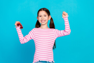 Sticker - Photo of cute positive small girl with ponytails dressed striped shirt hold binoculars clenching fist isolated on blue color background